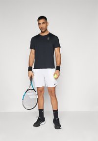 Svart trenings-T-skjorte med logo, hvite shorts, svarte sko og håndleddstrikker. Holder en tenniskølle og en grønn tennisball.