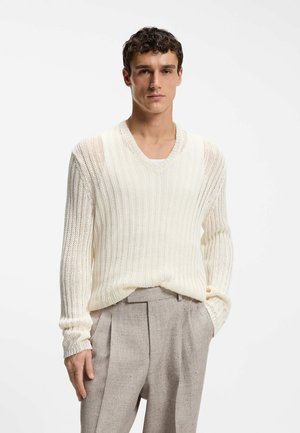 Joven con un suéter de punto color crema con cuello en V y pantalones plisados beige claro, de pie con una mano en el bolsillo contra un fondo liso.