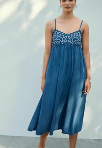 Denim midi-klänning med spaghetti-straps, med vit abstrakt broderi vid midjan. Klänningen har en lös, flödande form och en blå nyans.