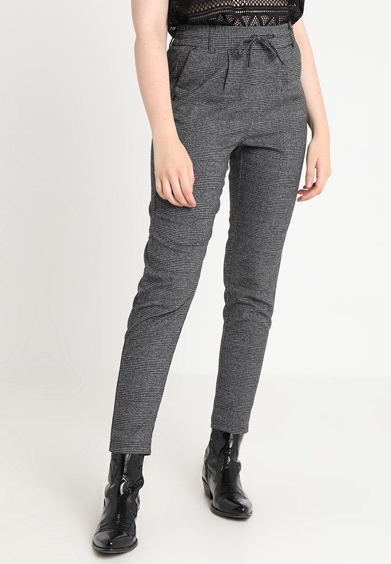 Femme portant un pantalon fuselé à carreaux gris avec une ceinture nouée à la taille et des bottines noires brillantes, debout devant un fond uni.