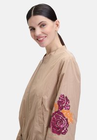 Beige bomberjack met een rits, voorzien van bloemige paillettenborduursel in roze en oranje op de linkermouw, een gladde textuur en een ontspannen pasvorm.