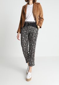 Bruine suède motorjack, witte T-shirt, zwarte bloemenbroek, witte sneakers met espadrille zool. Slim fit, verstelbare tailleband, enkel lengte.