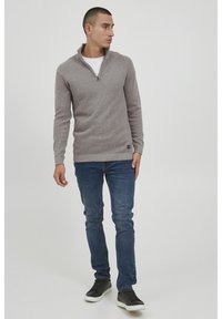 Pull en maille gris clair avec un col zippé, associé à un jean en denim bleu et des baskets noires. La texture est à motif et la coupe est décontractée.