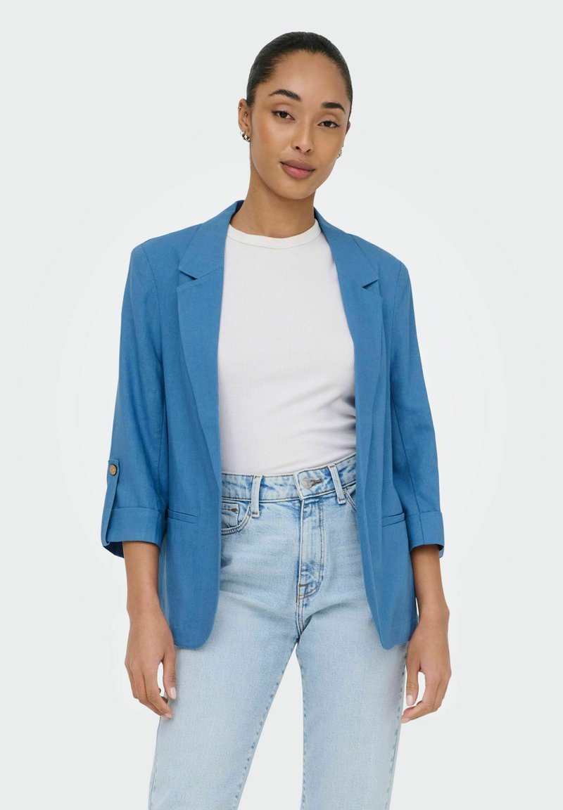 Femme portant un blazer bleu aux manches retroussées, un top blanc côtelé et un jean bleu clair taille haute sur un fond uni.