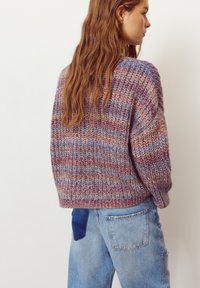 Pulover tricotat în nuanțe de mov, roz și albastru, cu un model texturat. Croială lejeră, mâneci largi, asortat cu blugi albaștri deschiși.