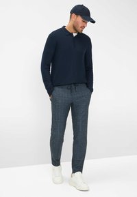 Marineblauwe lange mouwen polo, geruite donkergrijze broek met trekkoord, en witte sneakers. Gladde stof, getailleerde pasvorm, subtiele textuur.