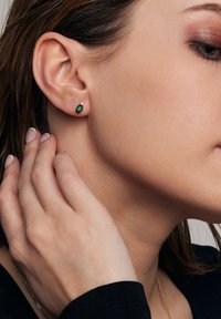 Kolczyki w kształcie szmaragdu z zielonym kamieniem, otoczone małymi białymi akcentami, osadzone w srebrze lub metalu w kolorze srebrnym. Noszone na uchu.