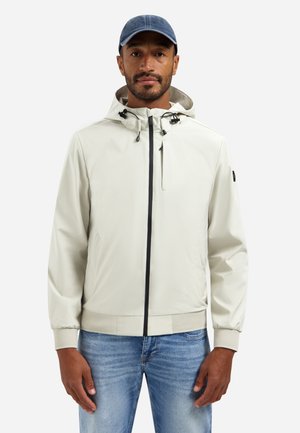 Man draagt lichtbeige capuchonjack met zwarte rits, blauwe spijkerbroek en donkerblauwe pet, staand tegen een effen witte achtergrond.