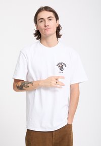Jeune homme aux cheveux mi-longs portant un T-shirt blanc Volcom, un pantalon marron et montrant un tatouage sur son avant-bras gauche.