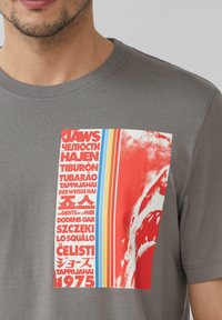 Grijze T-shirt met een grote grafische print in rood, blauw en wit. Het ontwerp bevat tekst in meerdere talen en een visueel element van een haai.