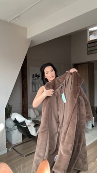 Καφέ παλτό από συνθετική γούνα που κρατιέται, με απαλή υφή και φαρδύ γιακά. Περιλαμβάνει ετικέτα και παρουσιάζεται με ουδέτερο εσωτερικό φόντο.