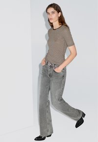 Pruhovaná hnědo-beige obepnutá košile v kombinaci s vysokými šedými džíny, doplněná černými podpatky na světlém pozadí.