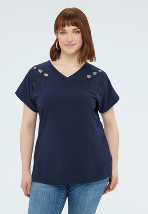 Donna con capelli rossi fino alle spalle che indossa orecchini a cerchio dorati, una maglietta blu navy a maniche corte con scollo a V e dettagli ritagliati, e jeans blu.