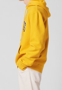 Sweat à capuche jaune en mélange de coton avec une poche avant, un design à enfiler, une capuche avec cordon de serrage et un texte noir contrastant sur le devant.