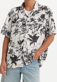 Camisetta a maniche corte con colletto in stampa floreale bianco e nero, caratterizzata da disegni di ibischi e palme, con bottoni e taschino sul petto.