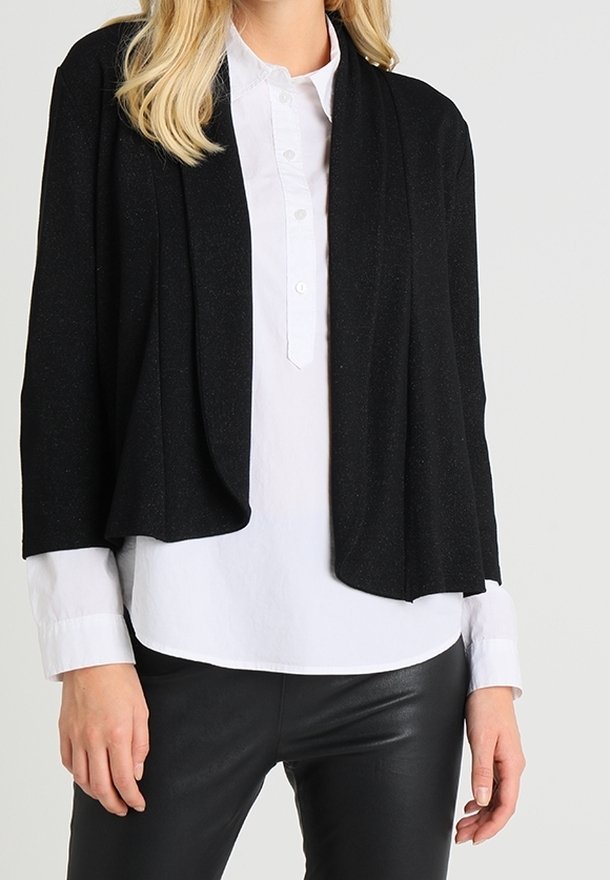 Svart cardigan med draperat framdesign, tillverkad av lättviktsmaterial. Bärs över en vit skjorta med knappar och kombineras med svarta läderbyxor.