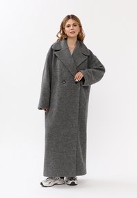 Cappotto grigio in misto lana con ampio revers, chiusura doppiopetto e vestibilità over, abbinato a sneaker sportive chiare.