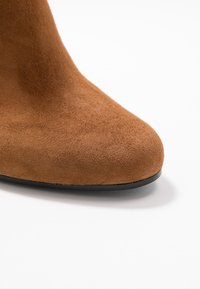 Brun ruskindsankelstøvle med en rundet tåsko, glat tekstur og en sort gummisål, der fremhæver det sømløse design.