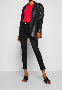 Blazer de cuero negro sobre un suéter rojo con cordones, combinado con jeans skinny negros y mulas negras de tacón con tachuelas plateadas.