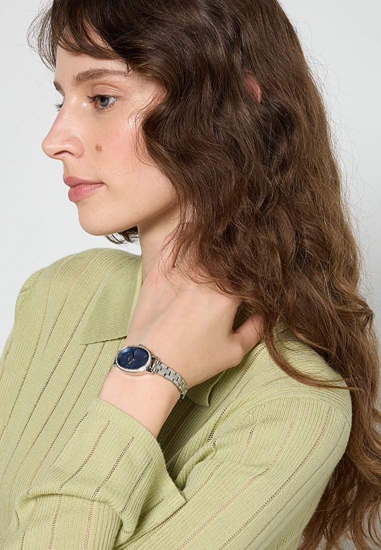 Montre en argent avec un cadran bleu rond, bracelet en métal à maillons, portée sur un poignet, complétée par un pull en maille texturée de couleur vert clair.