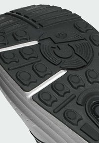 Suela de zapato de goma con una superficie texturizada negra que presenta patrones circulares y hexagonales, una línea de media suela blanca y un detalle de logo.