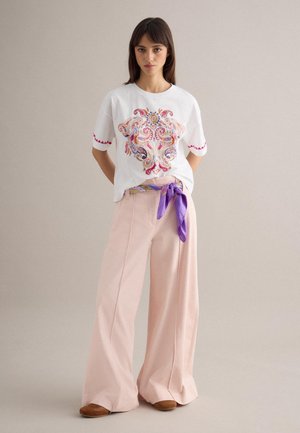 Jonge vrouw draagt een wit geborduurd T-shirt, wijde lichtroze broek, bruine schoenen en een paarse sjaal/broekriem met patroon, staand tegen een neutrale muur.