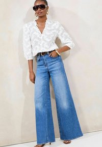 Weißes, transparentes Blusenshirt mit floralen Stickereien, kombiniert mit hoch taillierten, weit geschnittenen blauen Jeans. Ein schwarzer Gürtel und übergroße Sonnenbrillen unterstreichen den Look.