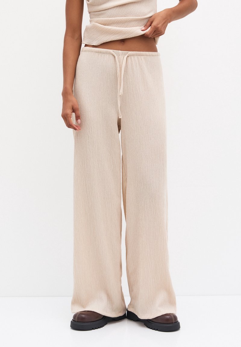 Beżowe spodnie o szerokich nogawkach wykonane z teksturowanego materiału, z elastycznym pasem i luźnym krojem, zestawione z brązowym obuwiem.