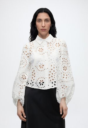 Donna con lunghi capelli neri che indossa una camicetta bianca floreale con ritagli e maniche a sbuffo e una gonna nera, rivolta verso la fotocamera su uno sfondo semplice.