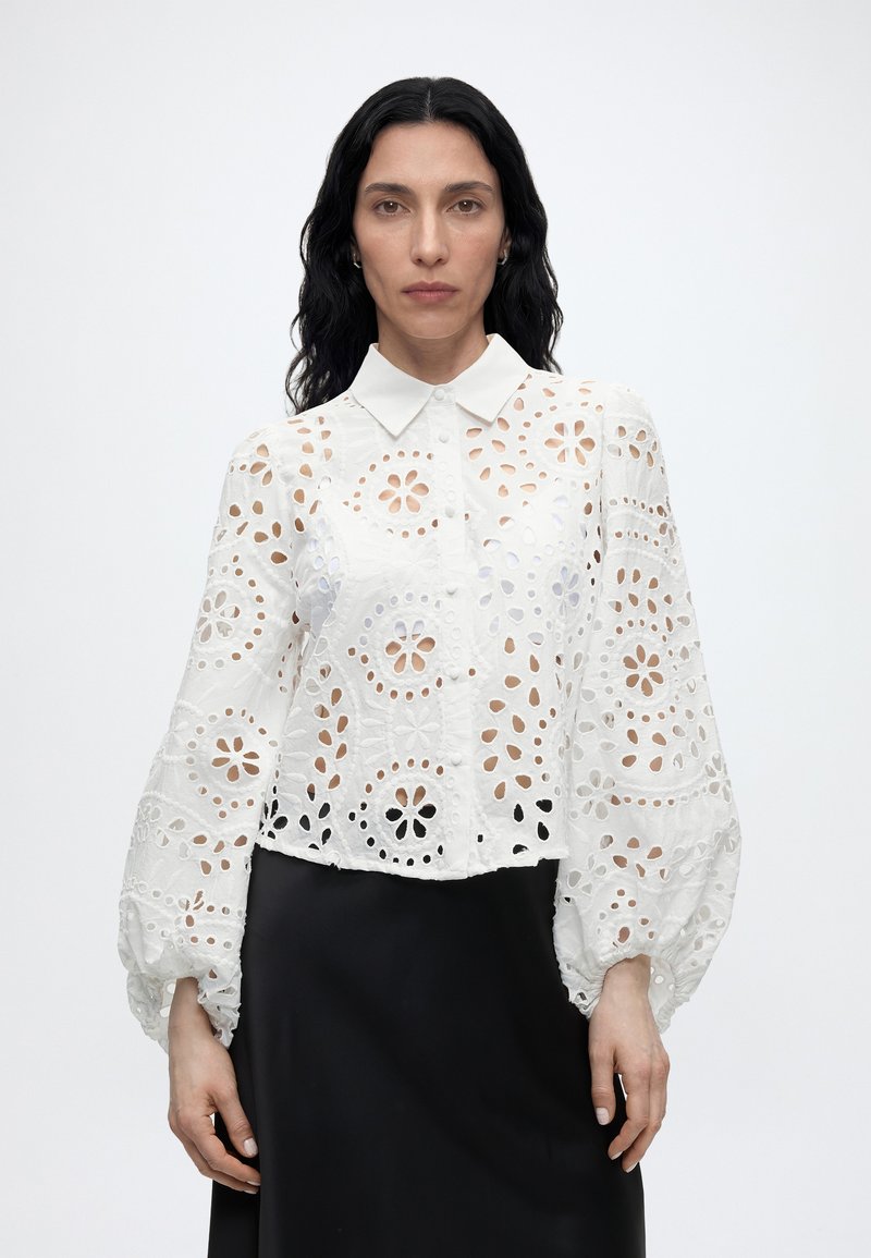 Γυναίκα με μακριά μαύρα μαλλιά, που φορεί μια λευκή μπλούζα με λουλουδάτα κοψίματα και φουσκωτά μανίκια και μια μαύρη φούστα, που κοιτάζει μπροστά σε απλό φόντο.