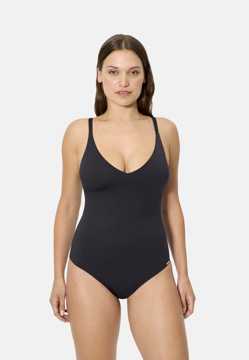 Mujer con cabello largo y castaño vistiendo un traje de baño negro de una pieza con tirantes delgados, de pie frente a un fondo claro y liso.