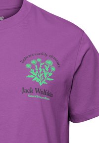 T-shirt violet avec un motif floral vert et le texte "Embrace earthly elements" au-dessus et "Jack Wolfskin Natural living wellness" en dessous.