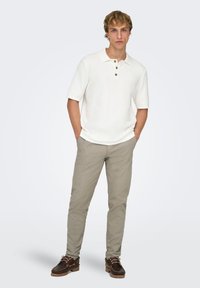 Polo à manches courtes blanches avec trois boutons, associé à un pantalon beige clair et des chaussures marron foncé avec des accents en lacets.