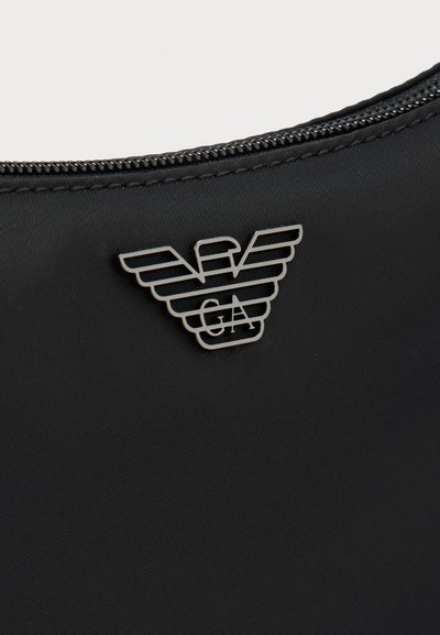 Κοντινό πλάνο μαύρου υφάσματος με ασημένιο μεταλλικό φερμουάρ και ασημένιο λογότυπο σε σχήμα αετού με τα γράμματα E, A και G.