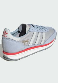 Gray suede and mesh sneaker featuring three white stripes, red accents on the sole, and a textured tongue. Gold logo detail on the side.