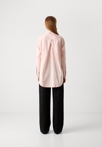 Kvinna med långt rakt hår som bär en lös ljusrosa skjorta med knappar och vida svarta byxor, stående vänd mot en helt vit vägg.