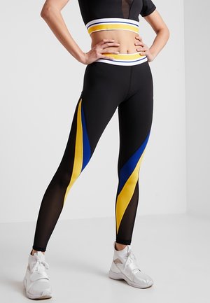 Persona che indossa leggings neri con strisce diagonali gialle e blu, un crop top abbinato e sneakers bianche, in piedi con le mani sui fianchi.