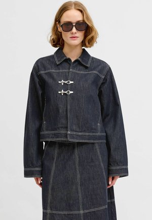 Kobieta ubrana w ciemną dżinsową kurtkę ze srebrnymi klamrami i pasującą spódnicę, uzupełniona dużymi czarnymi okularami przeciwsłonecznymi, stojąca na jednolitym tle.