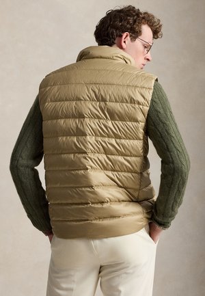 Homme aux cheveux bouclés portant des lunettes, un gilet matelassé marron clair par-dessus un pull en maille torsadée verte, et un pantalon couleur crème avec les mains dans les poches.