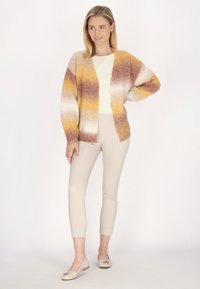 Cardigan lavorato a maglia in sfumature di giallo, arancione e marrone; design aperto davanti. Abbinato a pantaloni skinny beige e ballerine beige chiaro.