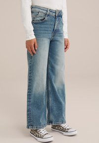 Široke modre kavbojke z obledelim dizajnom, petimi žepi in gumbom za zapiranje, v kombinaciji z škornji z leopardjim vzorcem.