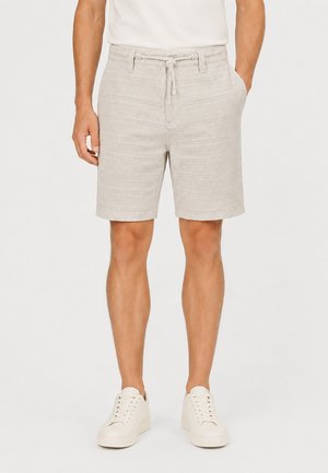 Mann trägt hellbeige strukturierte Shorts mit Kordelzug und Seitentaschen, weißes T-Shirt und weiße Sneaker, steht vor einem schlichten Hintergrund.