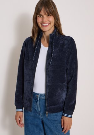 Femme souriante portant une veste zippée bleu marine avec des poignets rayés sur une chemise blanche et un jean bleu devant un fond uni.