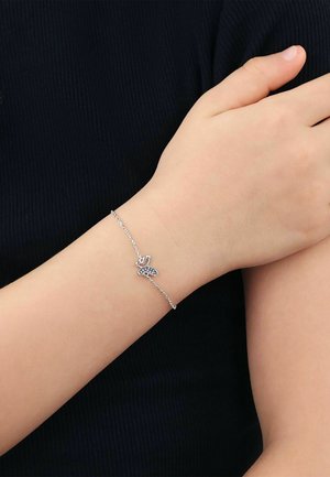 Feines silbernes Kettenarmband mit einem Schmetterlingsanhänger am Handgelenk einer Person vor einem dunklen, gerippten Stoffhintergrund.