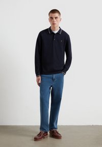 Marineblaues Langarm-Polo-Shirt mit weißen und roten Kragenakzenten, kombiniert mit hellblauen Jeans und braunen Schnürschuhen. Einfaches, lässiges Outfit.