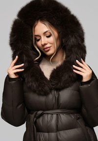 Cappotto imbottito nero con un ampio cappuccio in pelliccia marrone. Presenta una vita con cintura, texture trapuntata e maniche lisce.