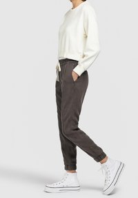 Persoon in een witte langemouwen shirt, donkergrijze joggingbroek met trekkoord en witte hoge sneakers, staand in profiel tegen een grijze achtergrond.