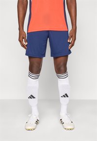 Άνδρας αθλητής που φοράει μπλε σκούρα σορτς, λευκές ποδοσφαιρικές κάλτσες με μαύρες ρίγες και άσπρα ποδοσφαιρικά παπούτσια, στέκεται μπροστά σε ουδέτερο φόντο.