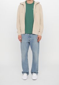 Sudadera con cremallera beige sobre una camiseta verde, combinada con jeans de mezclilla azul claro y zapatillas blancas. La sudadera cuenta con un cordón y bolsillos.