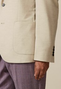 Blazer de herringbone bej deschis cu un buzunar, trei butoane negre pe manșetă, asortat cu pantaloni cu carouri mov. Accent pe textură și motive.